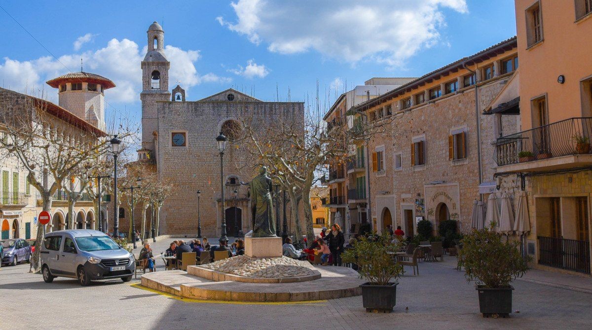 Town square with a church, statue, and people sitting at outdoor tables on a sunny day.