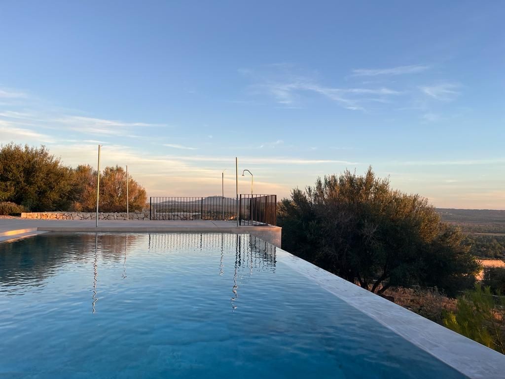Infinity pool overlooking a scenic landscape with trees and distant hills under a clear blue sky at sunset.